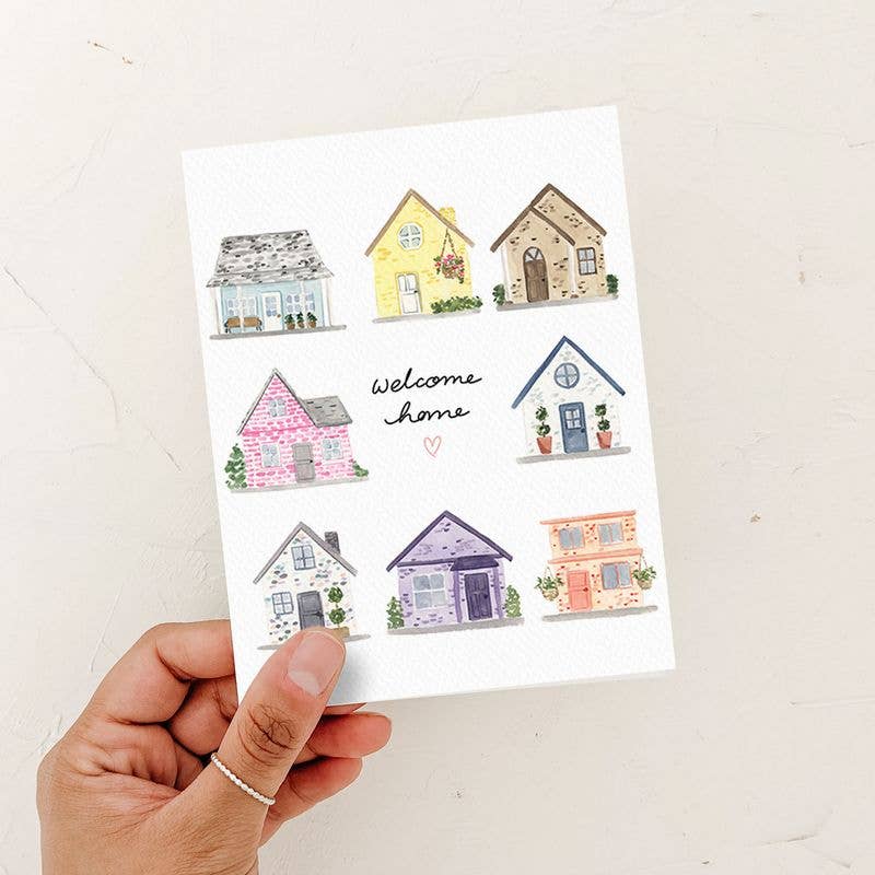 A person’s hand holding a watercolor "Welcome Home" greeting card featuring a grid of eight colorful houses against a textured white background.