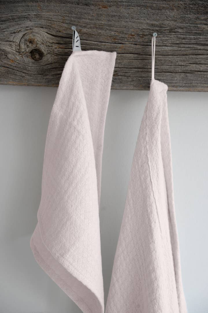 A light blush wash cloth and a matching hand towel hangs on a wooden rod against a white wall. 