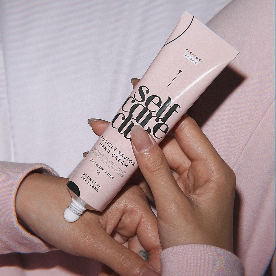 A person's hands are shown squeezing a pale pink tube of "Midnight Paloma Self Care Club Cuticle Savior Hand Cream," with a small dollop of white cream visible at the opening. The tube's label details "shea butter + rose" and "BRUNETTE THE LABEL" at the bottom. The background appears to be light pink clothing or fabric.