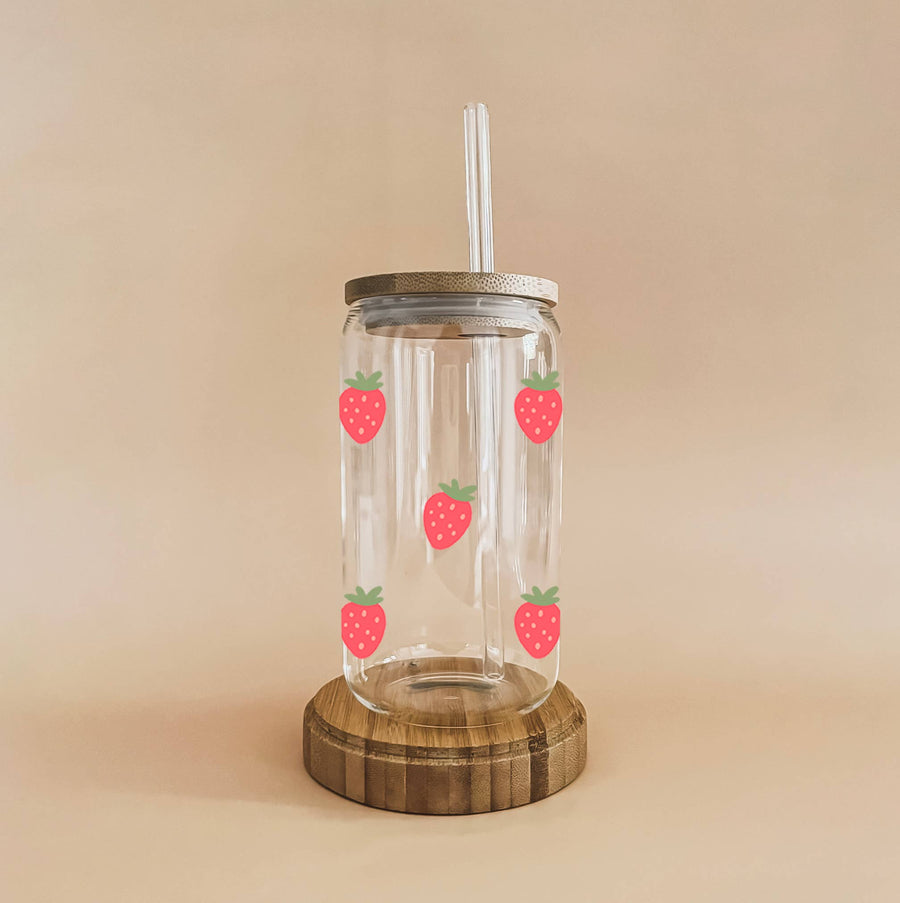 A clear glass cup with a bamboo lid and straw is decorated with red strawberry decals with green leaves and sits on a round wooden base.