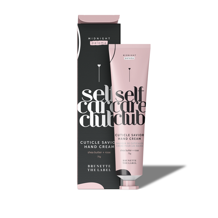 A pink tube of Midnight Paloma Self Care Club Cuticle Savior Hand Cream next to its black box. The tube and box have text in English and French, featuring 'shea butter + rose' and 'BRUNETTE THE LABEL.'