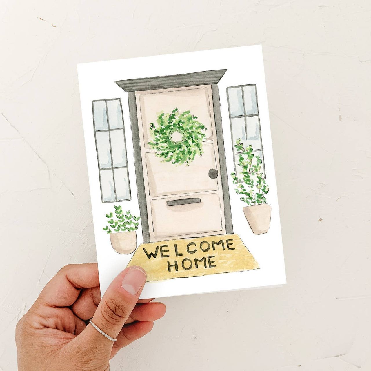 A person’s hand holding a watercolor "Welcome Home" greeting card featuring a front door with a wreath and potted plants against a textured white background.
