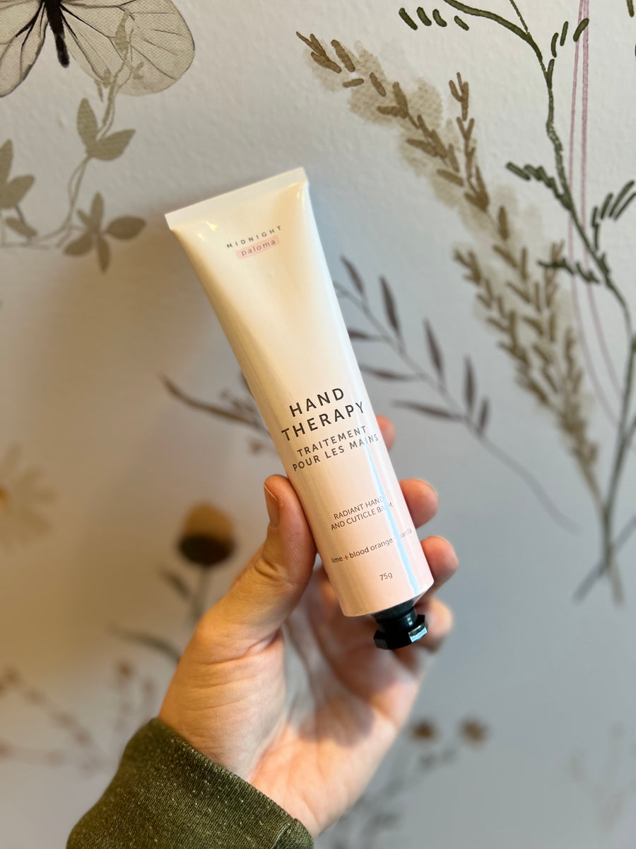 
A hand holds a light pink tube of "Midnight Paloma Hand Therapy" lotion with French text "TRAITEMENT POUR LES MAINS" and "RADIANT HAND AND CUTICLE BALM" visible, against a floral wallpaper background.