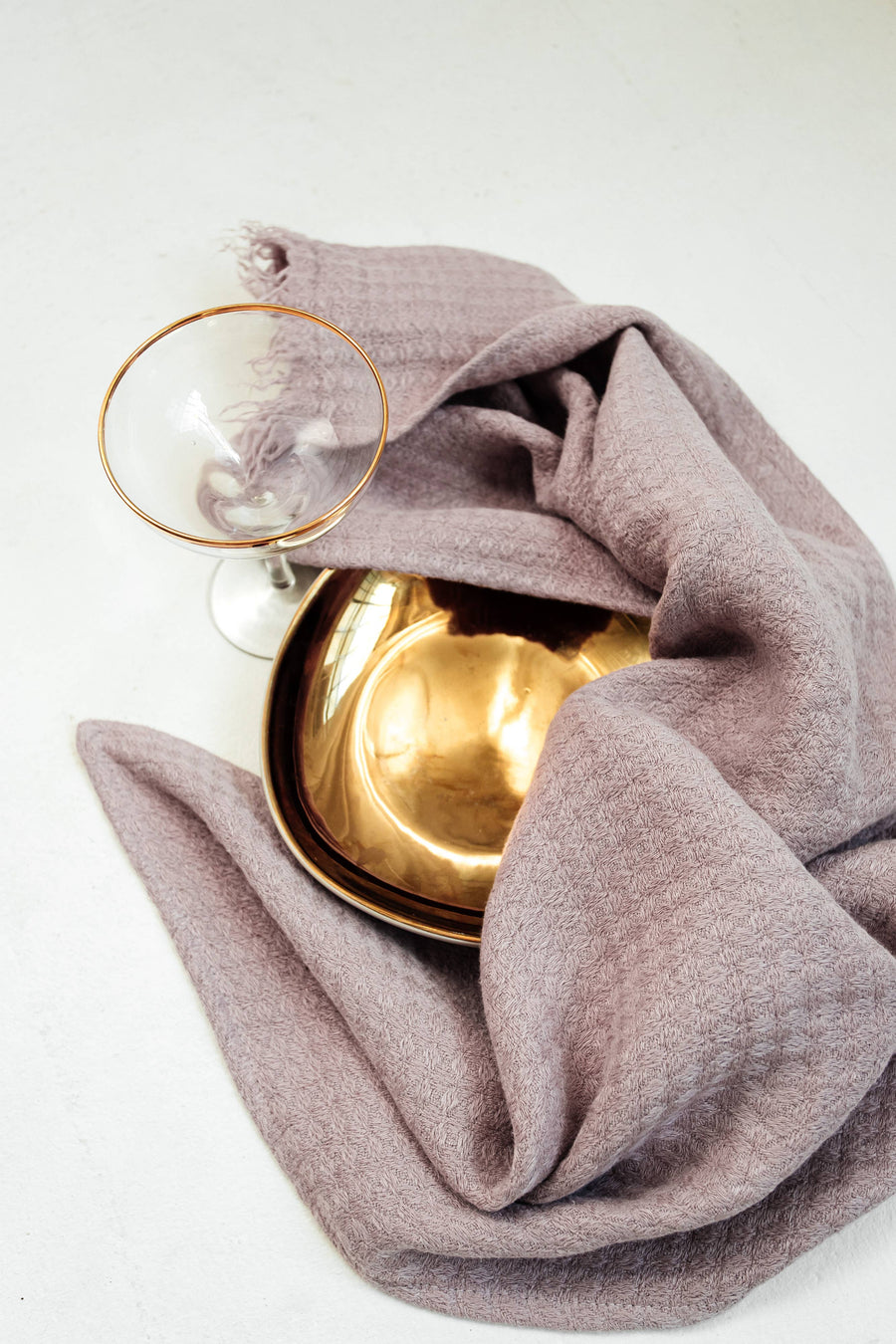 Gold bowl and glass with a lavender ask towel folded over the bowl on a white background