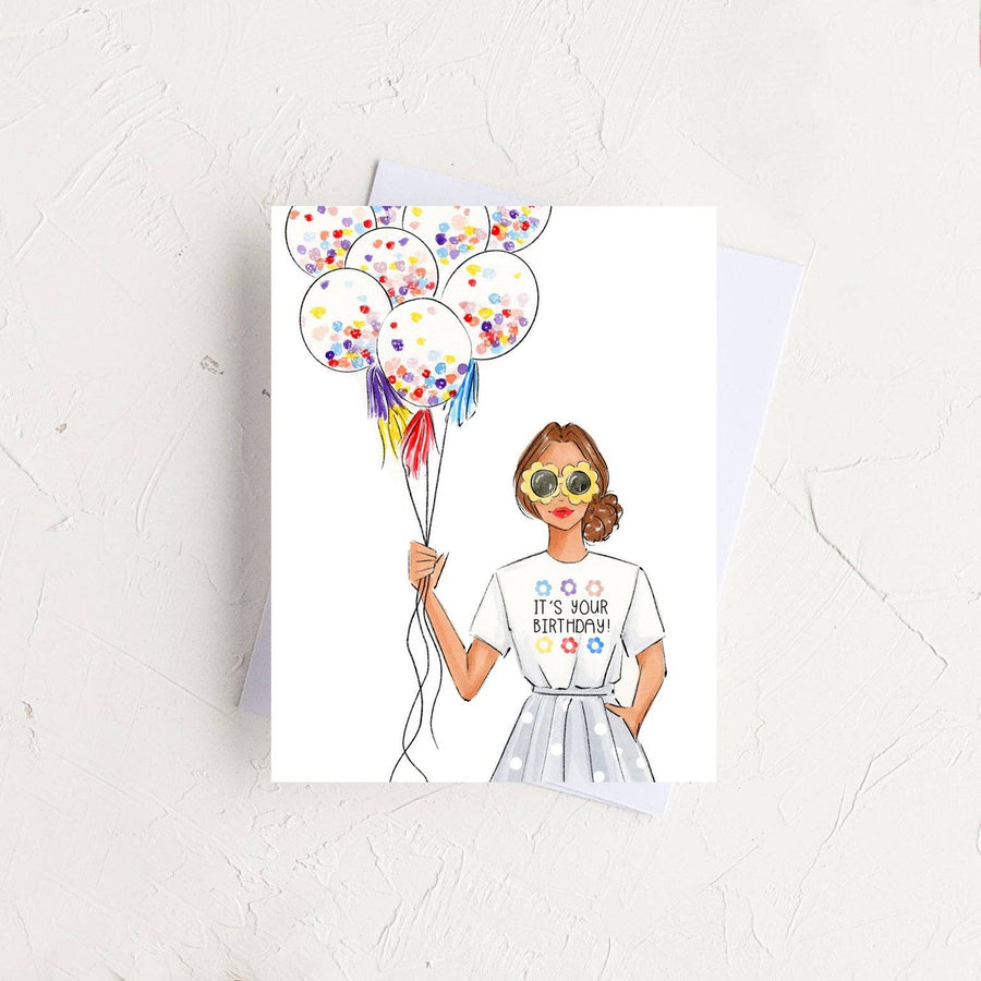 A watercolor "It's Your Birthday" card resting on a white envelope over a rustic, textured white surface, featuring an illustration of a fashionable woman holding confetti balloons.