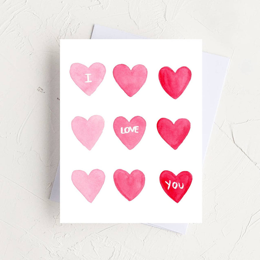 A watercolor card with a grid of nine pink and red hearts reading "I Love You," displayed on a white envelope over a rustic, textured white surface.