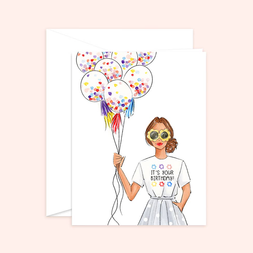 Watercolor birthday card and white envelope featuring an illustration of a fashionable woman in flower-shaped sunglasses holding a bunch of confetti balloons, wearing a shirt that says "IT'S YOUR BIRTHDAY!"
