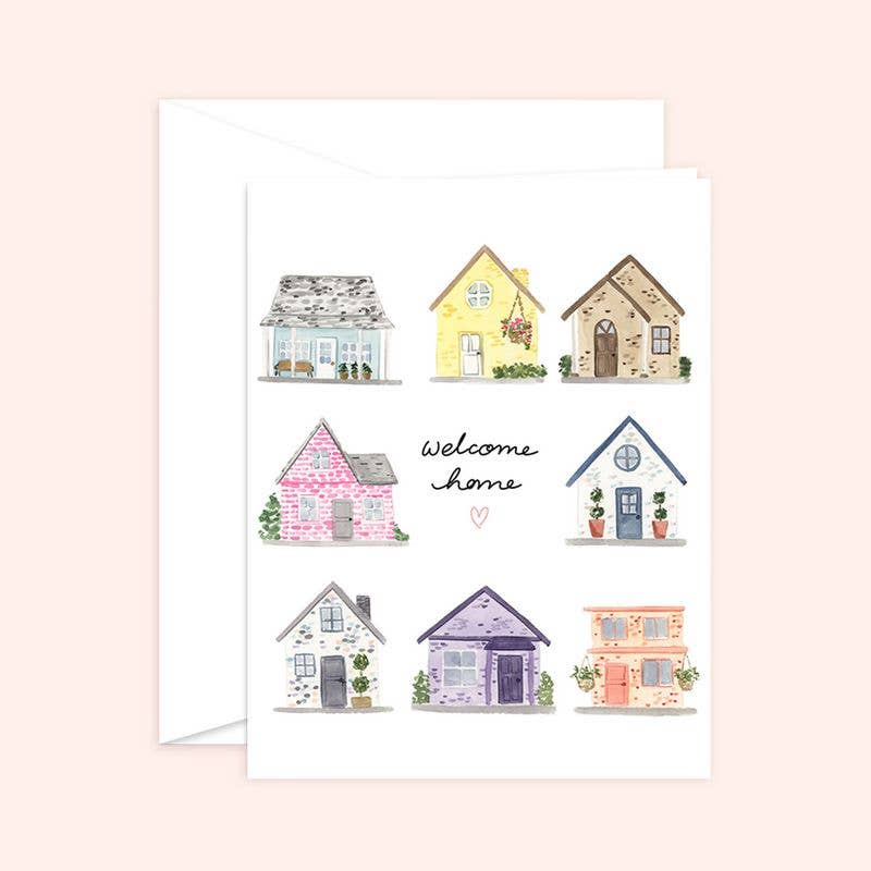 Watercolor greeting card and white envelope featuring a grid of eight unique, colorful houses surrounding the handwritten words "welcome home" with a small pink heart.