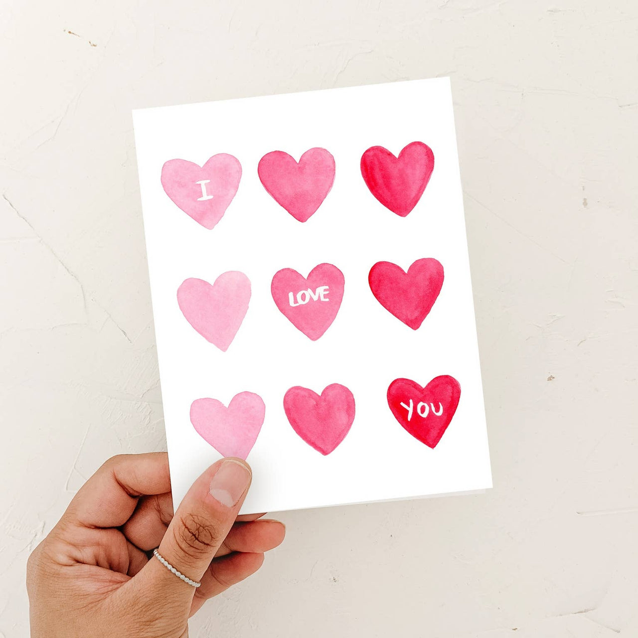 A person’s hand holding a watercolor "I Love You" card featuring a grid of pink and red hearts against a textured white background.