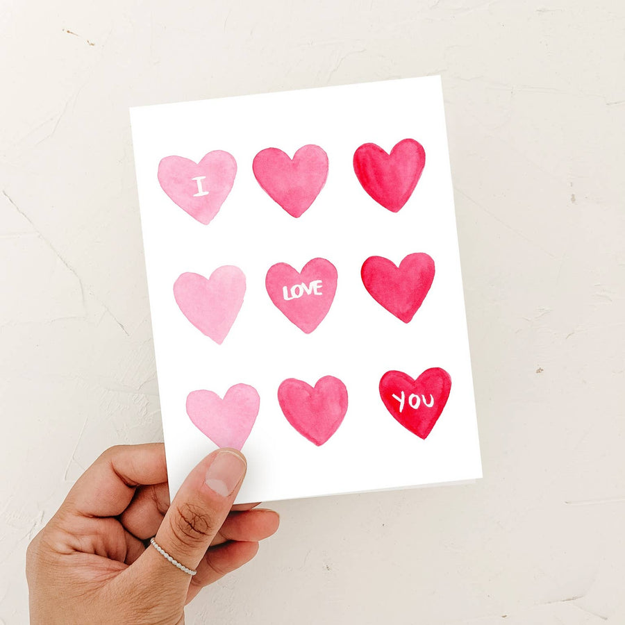 A person’s hand holding a watercolor "I Love You" card featuring a grid of pink and red hearts against a textured white background.
