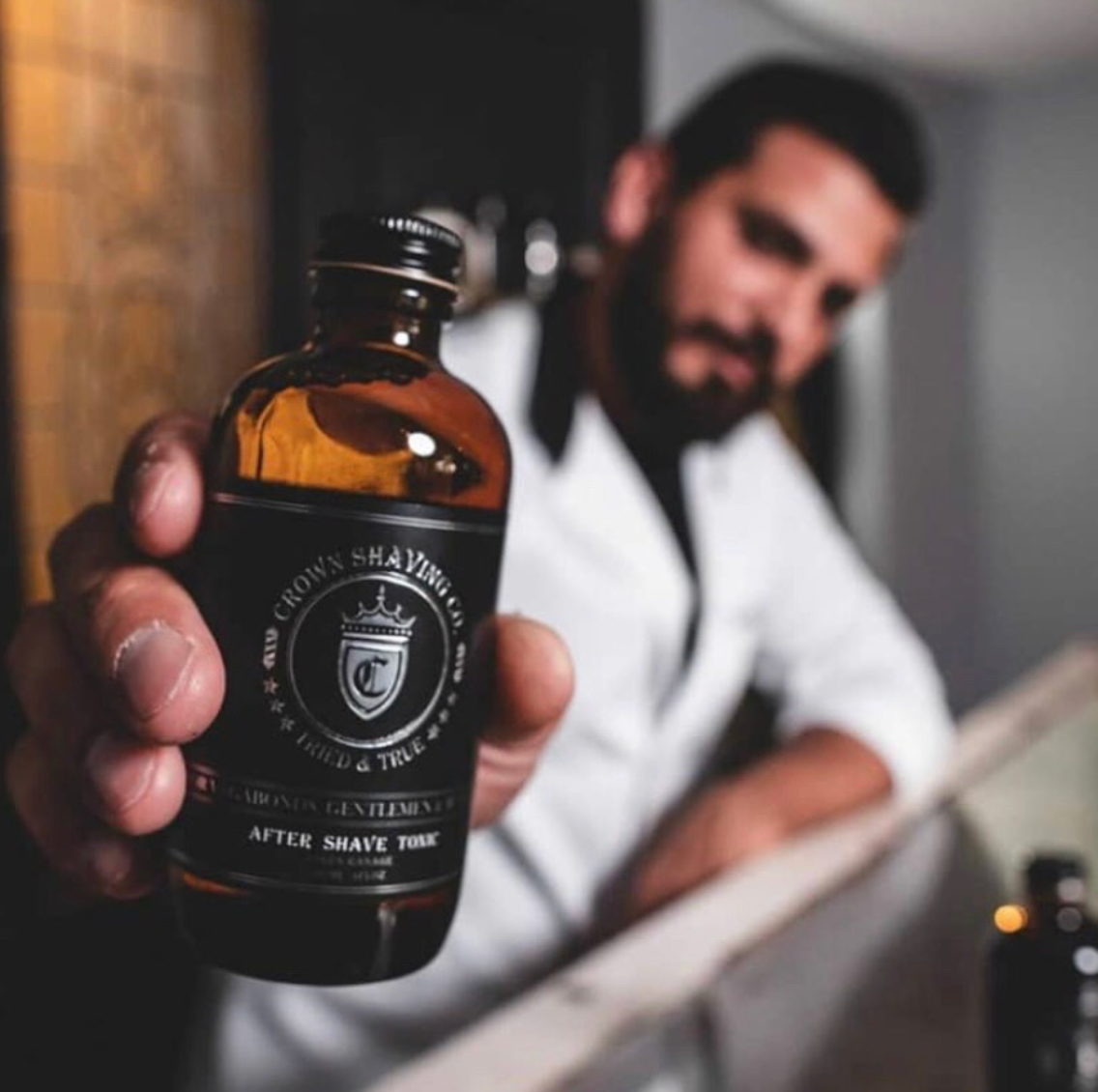 A person with a beard is holding out a brown glass bottle of Crown Shaving Co. After Shave Tonic towards the viewer. The label is clearly visible, featuring the brand's logo and the product name. The background is blurred, suggesting a barbershop setting.