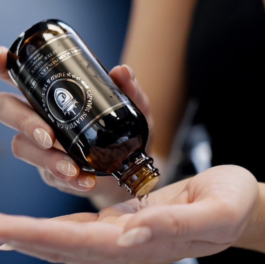 This close-up shot shows someone with manicured nails pouring Crown Shaving Co. After Shave Tonic from a brown glass bottle into their other hand. The label with the brand's logo and product name is partially visible on the wet bottle. The background is blurred.