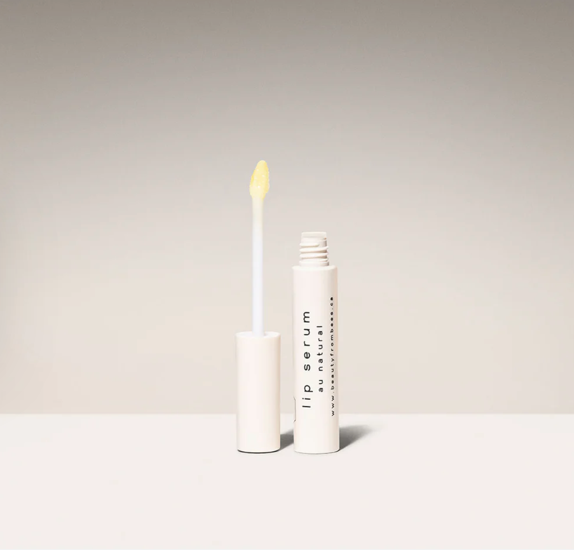 A white tube of "Lip Serum Au Natural" by Beauty from Bees, shown open with the applicator wand held upright to reveal the clear, glossy serum.