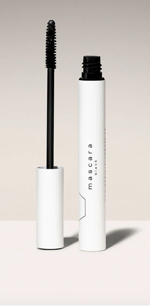 A white tube of "mascara black" with a black cap removed and the black mascara wand resting beside it. The tube has simple black lettering and a minimalist geometric design. The background is a soft, gradient beige.