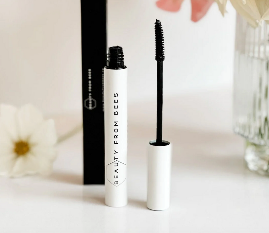 A white tube of "BEAUTY FROM BEES" mascara with a black cap removed and the black mascara wand resting beside it. The brand name is printed vertically on the tube. The mascara is positioned in front of its black packaging box and a blurred background with flowers
