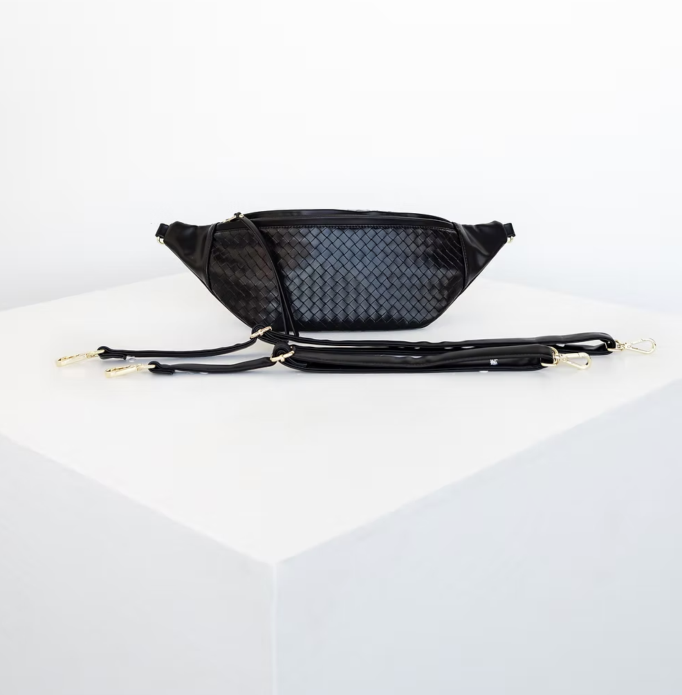 A black, woven-textured fanny pack sits on a white surface with two detachable straps lying in front of it.