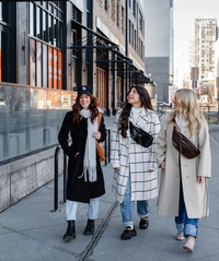 Thumbnail for hree women walk down a city sidewalk. The woman on the left wears a black coat, the one in the middle a white and black checkered coat with a black fanny pack, and the woman on the right a tan coat with a brown fanny pack.