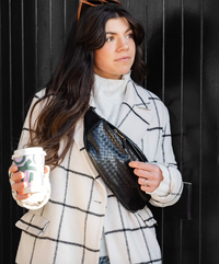Thumbnail for A woman in a white and black checkered coat holds a coffee cup and a black, woven-textured fanny pack worn crossbody.