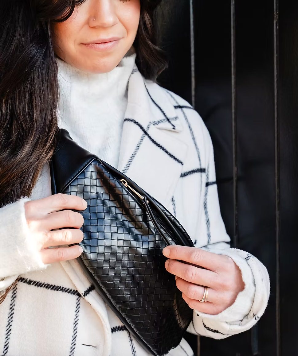 A close-up of a woman wearing a white sweater and a checkered coat as she opens a black, woven-textured fanny pack.