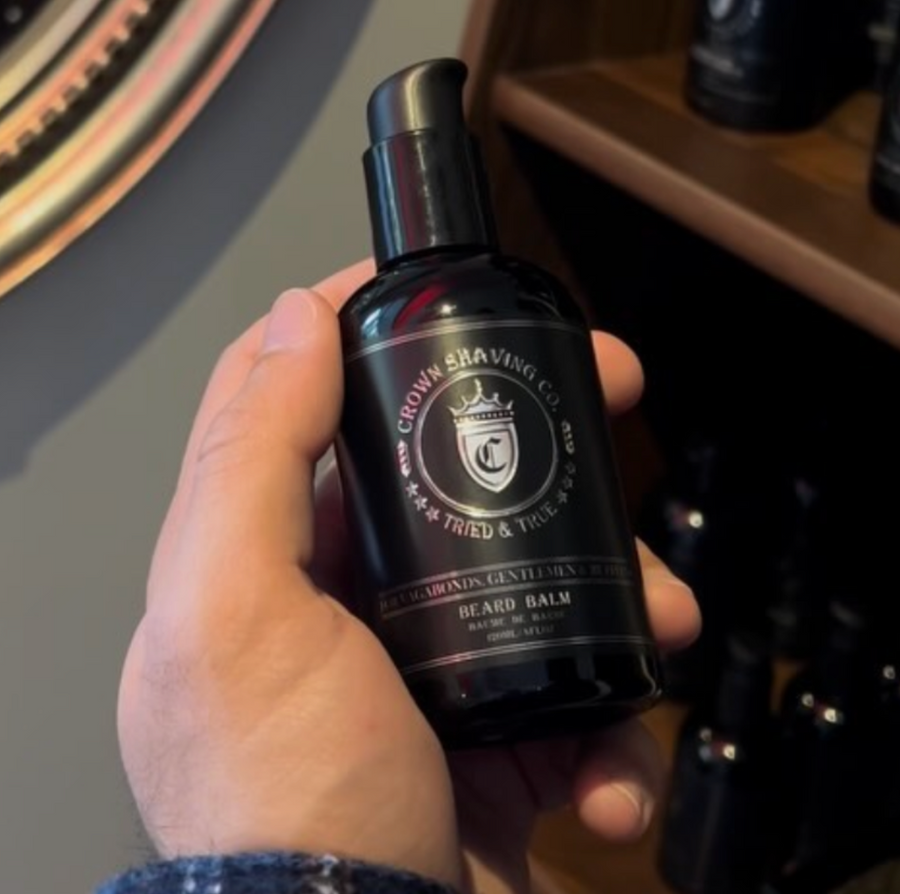 This image shows a black pump bottle of Crown Shaving Co. Beard Balm. The label features the brand's logo and the product name "BEARD BALM BAUME DE BARBE". The bottle is being held in a person's hand.