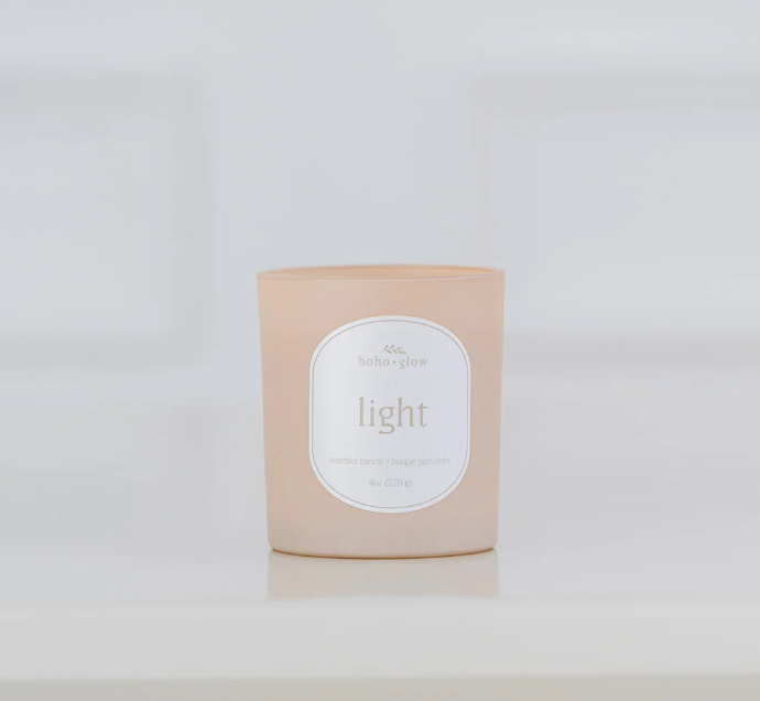 A frosted, light pinkish-beige candle in a glass jar sits on a white surface. The label reads "boho glow" and "light" with "8oz" and "scented candle" in smaller text below. The background is a blurry white tile.