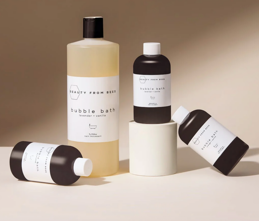 
A still life image featuring four bottles of "Beauty From Bees" bubble bath. One large, clear bottle of lavender vanilla scent stands upright. Two smaller, dark-colored bottles (also lavender vanilla) are lying on their sides in the foreground, and one smaller dark bottle is standing upright on a white cylindrical pedestal.