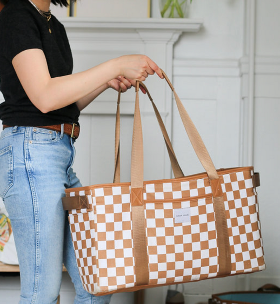 A person in a black shirt and jeans holds a large, brown and white checkered tote bag by its two straps.