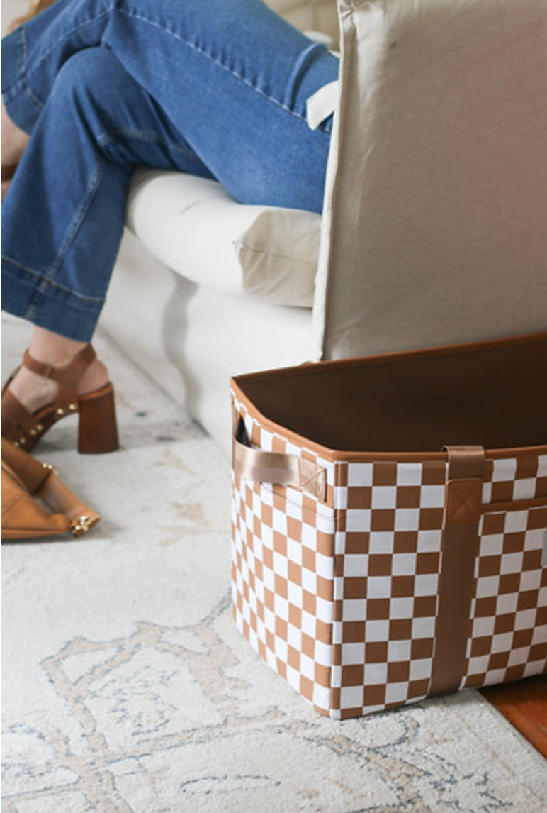 A brown and white checkered tote bag sits on a light-colored rug. In the background, a person wearing jeans and brown shoes with wooden heels sits on a white couch.