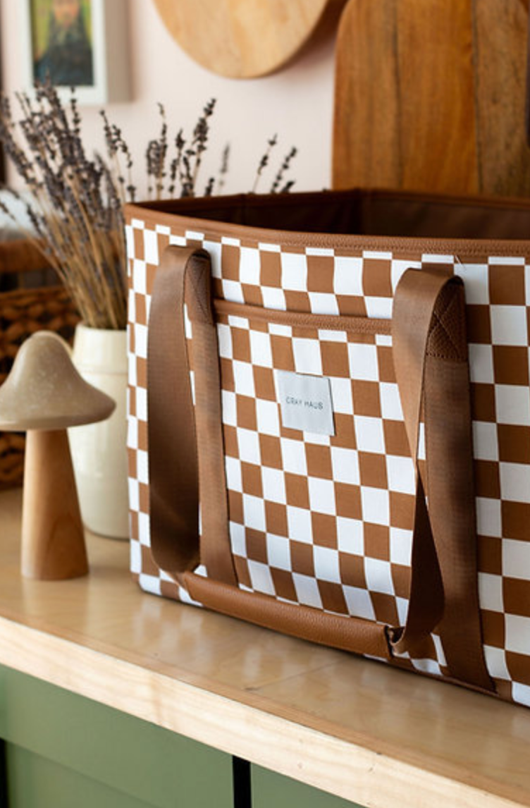 A close-up of a brown and white checkered tote bag with brown straps. The bag has a small logo patch on the front, and a wooden mushroom-shaped object is visible next to it on a counter.