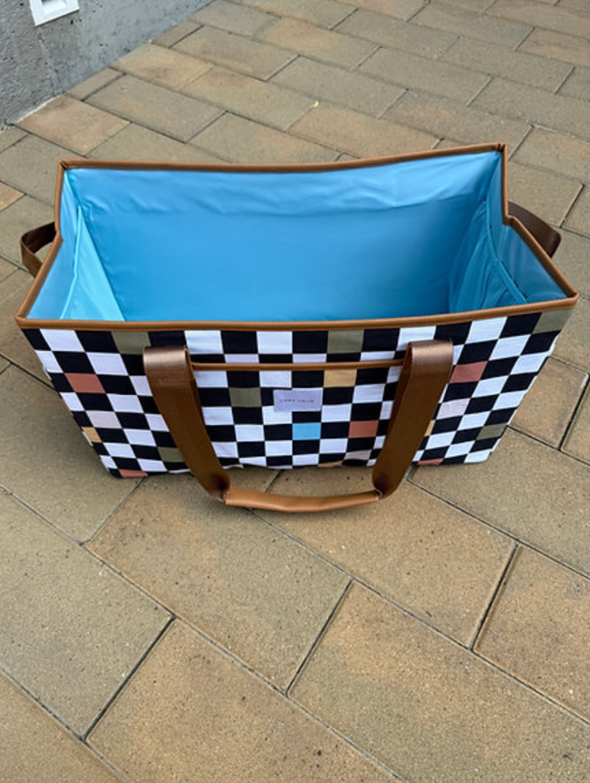 An open, rectangular fabric tote with a black and white checkerboard pattern and multicolored squares sits on a brick patio. The inside is lined with bright blue fabric, and the brown straps and handles are visible.