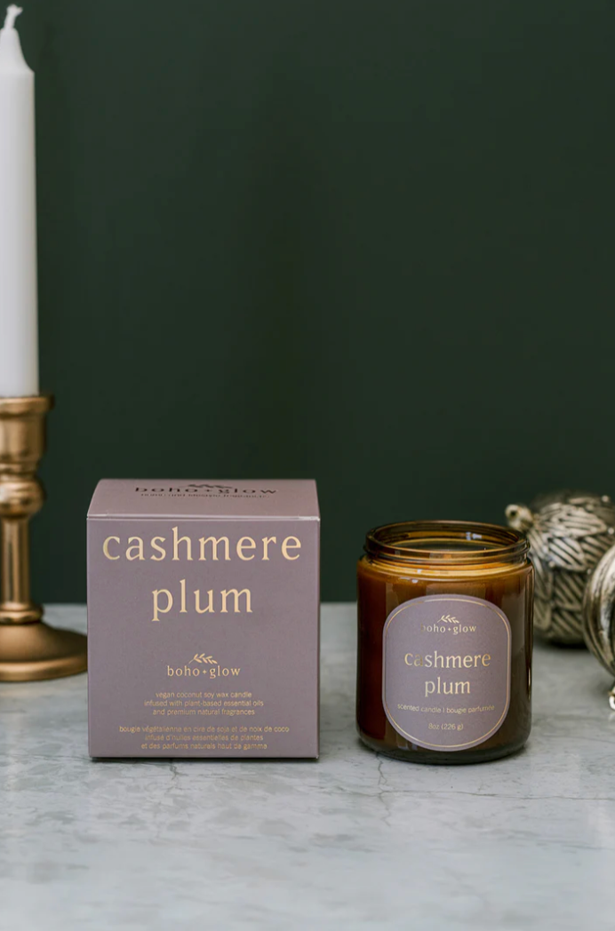 An amber jar candle and its square box, both labeled "cashmere plum" by Boho + Glow. The box is a dusty purple color, and a brass candle holder with a white taper candle is visible on the left against a dark green background.
