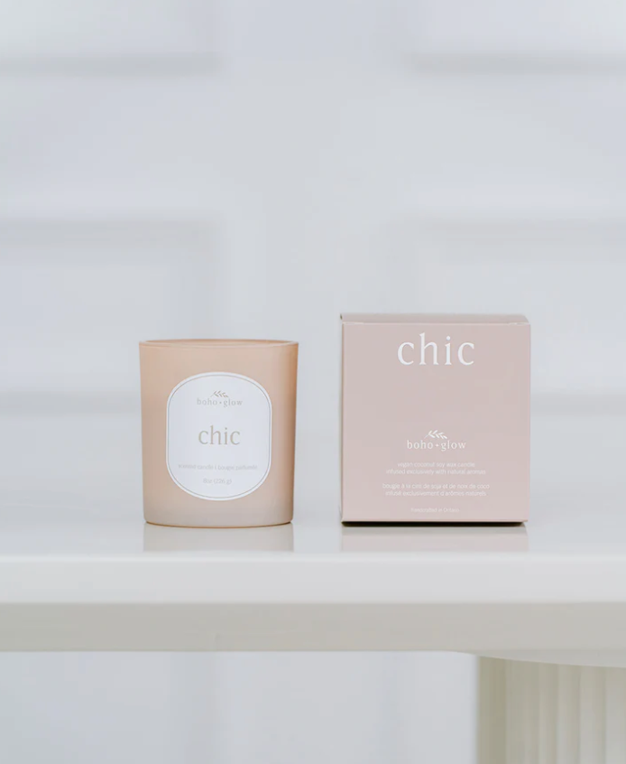 Beige candle labeled "chic" next to its matching box on a white shelf.