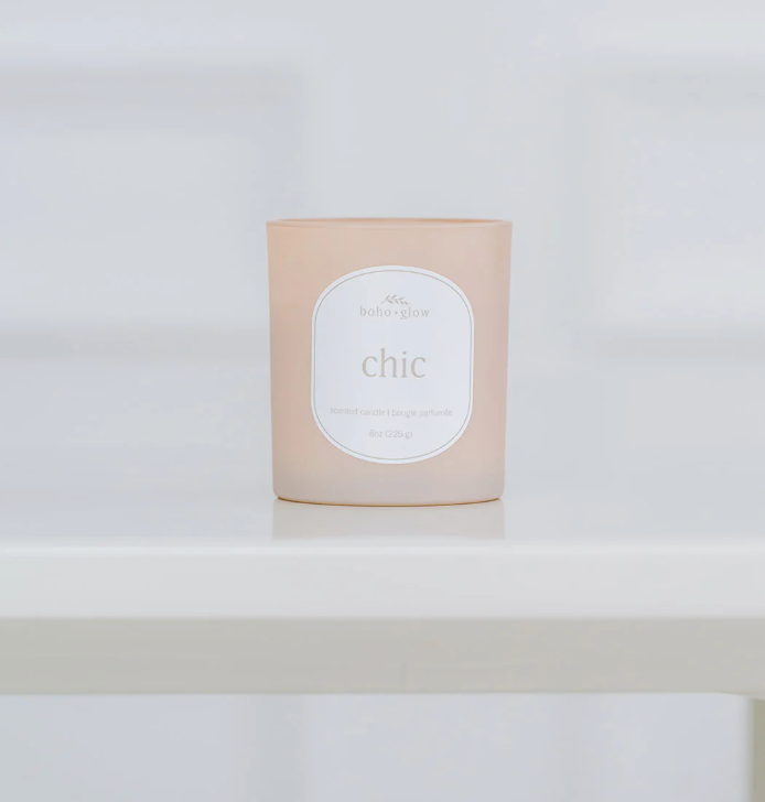 
A frosted, light pinkish-beige candle in a glass jar sits on a white surface. The label reads "boho glow" and "chic" with "8oz" and "scented candle" in smaller text below. The background is a blurry white tile.