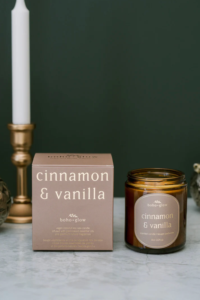 
A small amber jar candle and its matching tan box, both labeled "cinnamon & vanilla" by Boho + Glow, sit on a white marble surface. A tall white taper candle in a brass holder is partially visible on the left against a dark green background.