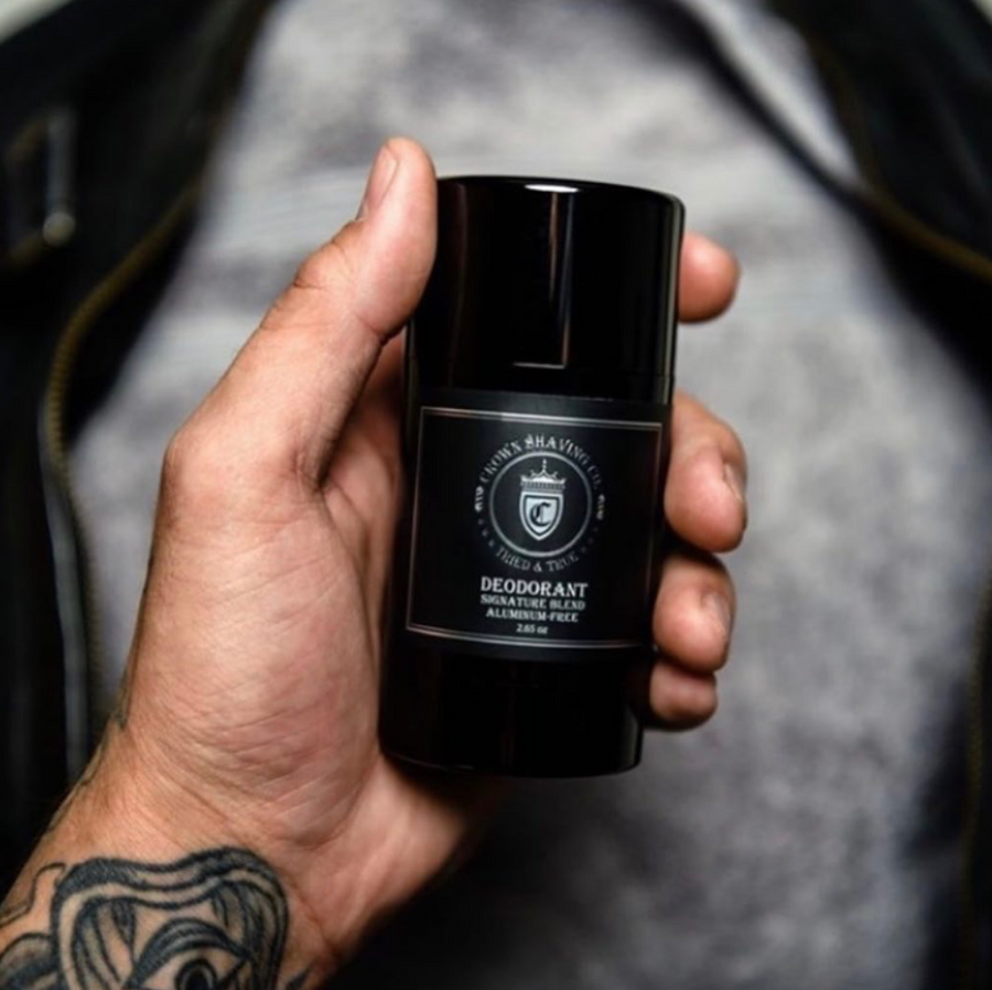 This close-up shot shows a tattooed hand holding a black container of Crown Shaving Co. Aluminum-Free Deodorant. The label is clearly visible, displaying the brand's logo and the product name. The background is slightly blurred, suggesting the person might be wearing a grey shirt and a black jacket.