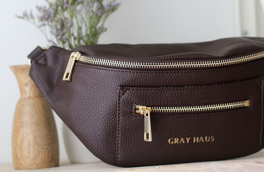 A close-up of a dark brown belt bag with gold zippers. The words "GRAY HAUS" are subtly printed below the smaller front zipper.