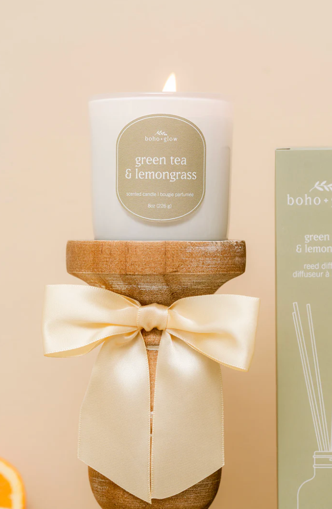 A white glass candle by Boho + Glow sits lit on a turned wooden pedestal. The sage green label reads "green tea & lemongrass" in white serif font. A large, cream-colored satin bow is tied around the pedestal, and a matching reed diffuser box is partially visible to the side against a soft, neutral background.