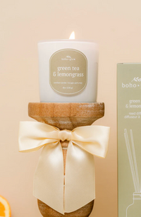 Thumbnail for A white glass candle by Boho + Glow sits lit on a turned wooden pedestal. The sage green label reads 