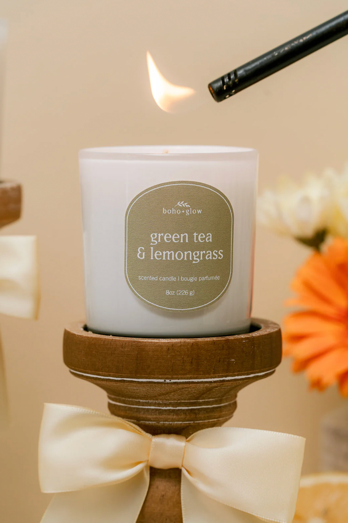 A close-up of a lit Green Tea & Lemongrass candle being ignited by a black candle lighter. The candle sits on a wooden stand decorated with a cream satin bow, with a vibrant orange gerbera daisy in soft focus in the background, evoking a fresh, botanical mood.