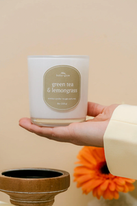 Thumbnail for A person’s hand holds the Green Tea & Lemongrass candle, showcasing its frosted glass and minimalist sage green label. In the background, an orange gerbera daisy and the corner of a wooden pedestal add warmth and color to the clean, modern composition.
