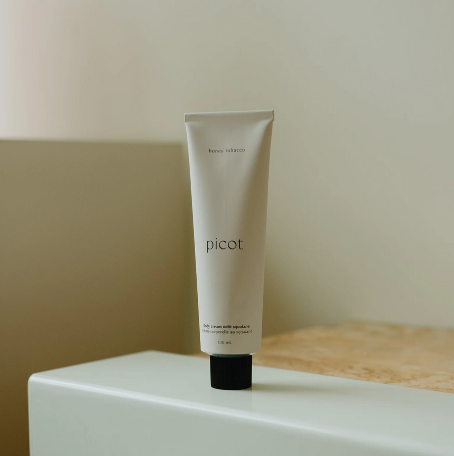 A simple, minimalist white tube of "picot honey tobacco" body cream with a black cap stands upright on a white and beige surface.