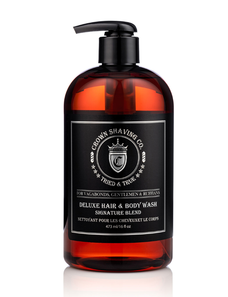 This image shows a clear bottle with an amber-colored liquid, identified by the label as Crown Shaving Co. Deluxe Hair & Body Wash. The black label features the brand's logo and the product name "DELUXE HAIR & BODY WASH SIGNATURE BLEND NETTOYANT POUR LES CHEVEUX ET LE CORPS." The bottle has a black pump dispenser.