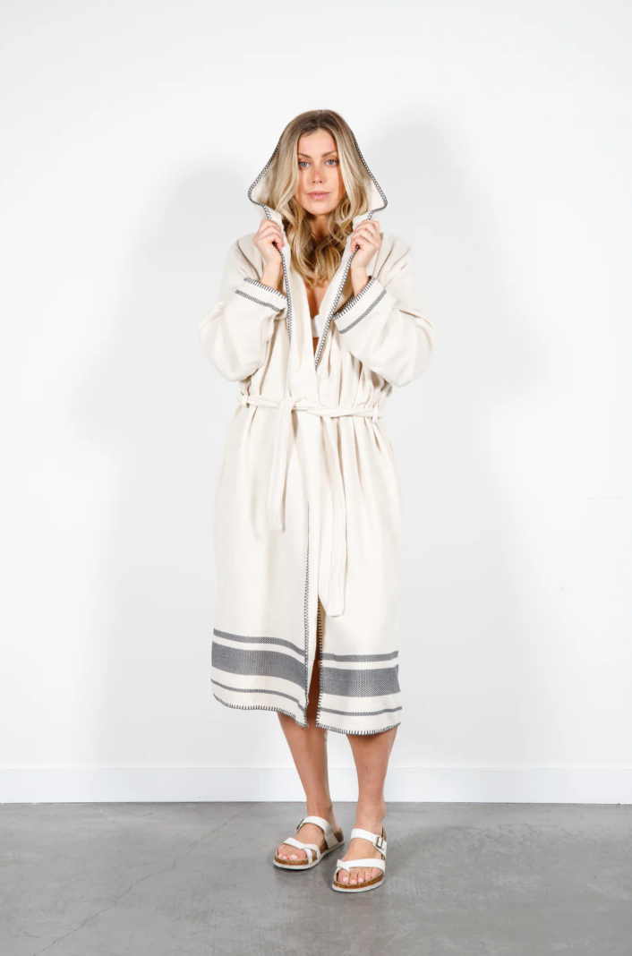 A person with long blonde hair models a long, hooded, off-white Turkish towel robe with grey stripes at the hem and cuffs. The robe is belted at the waist, and the model is wearing white sandals. They are standing against a plain white wall and a light grey concrete floor.