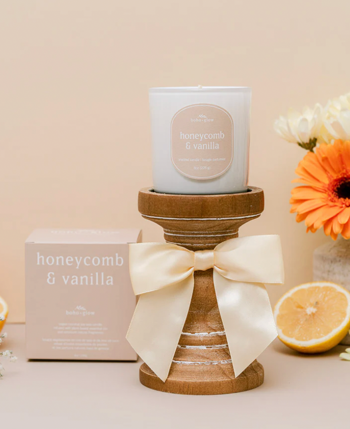 A white glass candle by Boho + Glow sits on a rustic wooden pedestal accented with a large, cream-colored satin bow. The tan label reads "honeycomb & vanilla" in white text. To the left, the matching tan gift box is visible, and to the right, a sliced lemon and an orange gerbera daisy add a warm, citrusy glow to the scene.