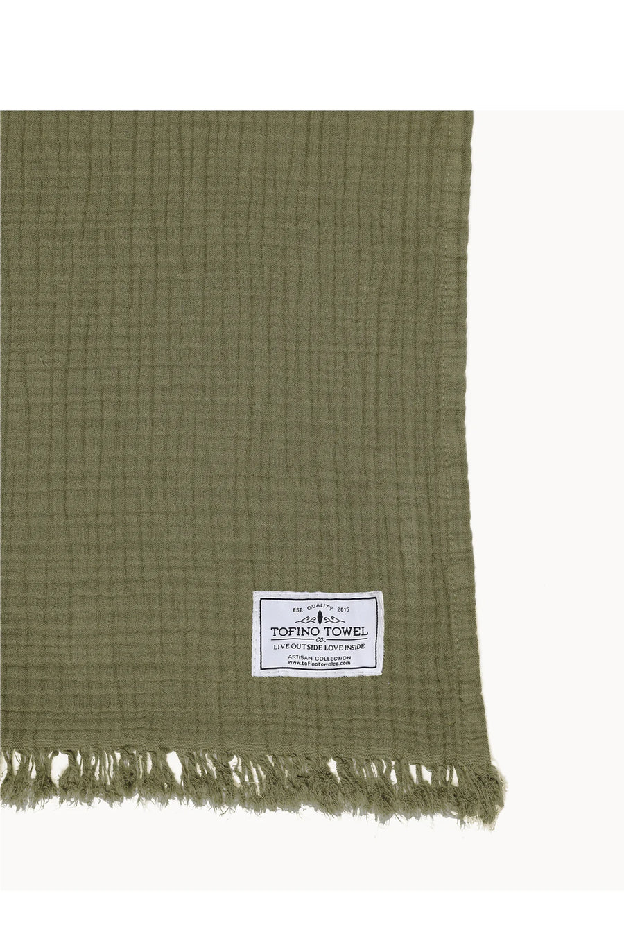 A close-up of a textured, olive green Tofino Towel with a fringed edge at the bottom. A cream-colored fabric label, stitched onto the towel, reads "TOFINO TOWEL" in bold letters, with smaller text and a logo above and below it.