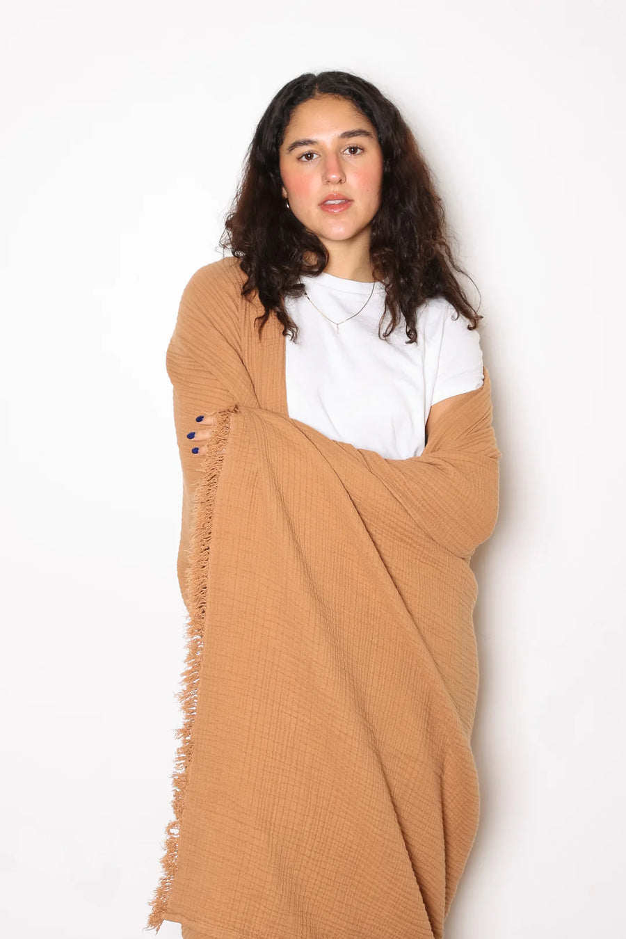 A young woman with long, dark curly hair stands against a white background, looking directly at the viewer. She is wearing a simple white t-shirt and a delicate silver necklace, and her fingernails are painted dark blue. She is wrapped in a large, textured, light brown or camel-colored throw blanket that has a fringed edge visible on her left side.