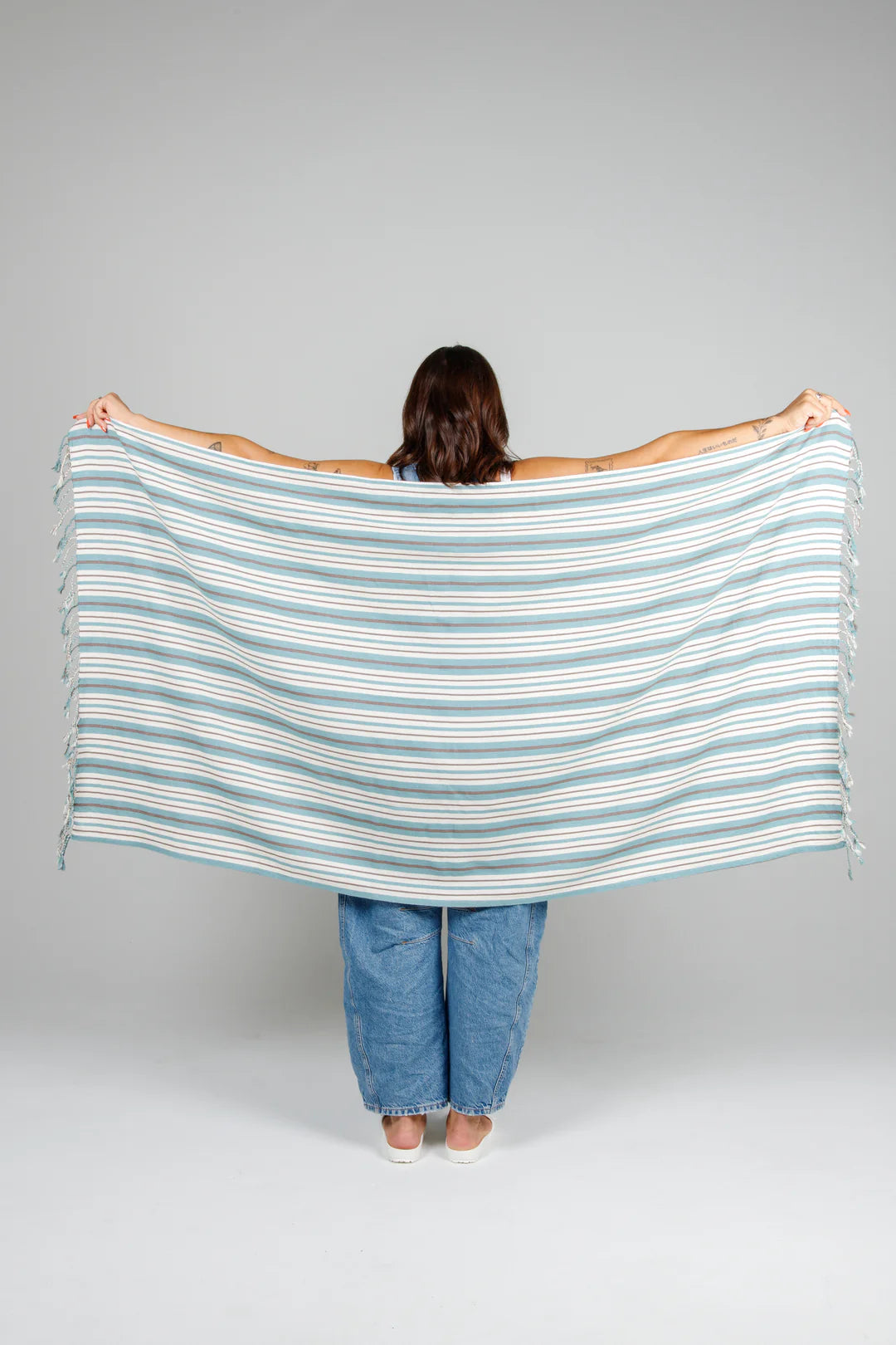 A woman holding the ocean-blue striped Mistral towel spread wide across her back, showcasing its full 160cm length and lightweight, flat-woven Turkish cotton texture.