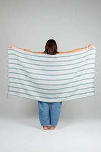 Thumbnail for A woman holding the ocean-blue striped Mistral towel spread wide across her back, showcasing its full 160cm length and lightweight, flat-woven Turkish cotton texture.