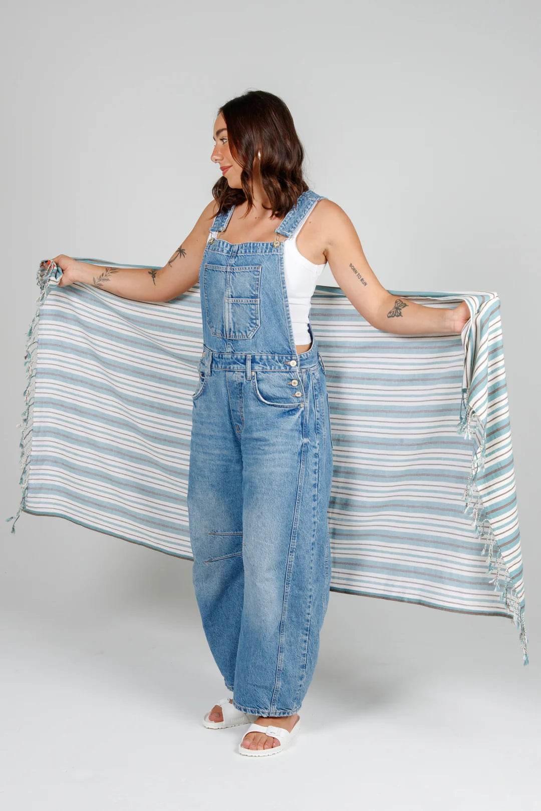 A woman in denim overalls and a white tank top holds the large, striped Mistral towel open in front of her. The towel features a pattern of ocean-blue, white, and thin grey horizontal stripes with twisted tassel fringe at the ends.
