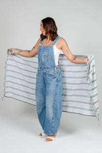 Thumbnail for A woman in denim overalls and a white tank top holds the large, striped Mistral towel open in front of her. The towel features a pattern of ocean-blue, white, and thin grey horizontal stripes with twisted tassel fringe at the ends.